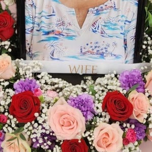 May include: A floral arrangement with pink, red, and white roses, purple carnations, and white baby's breath. The arrangement is in a heart shape and has a white ribbon with the word "WIFE" printed on it.