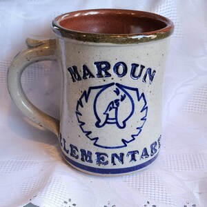 May include: A white ceramic mug with a blue and white design. The mug has a handle and says "Maroun Elementary" on the front.
