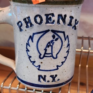 May include: A speckled ceramic mug with the word "PHOENIX" in raised blue letters. Below is a blue emblem and the letters "N.Y." The mug has a handle and a dark green rim.