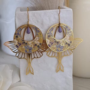 May include: A pair of gold earrings with a butterfly design. The earrings feature a circle with a floral pattern in shades of purple and gold, and a dangling butterfly shape. The earrings have a purple teardrop-shaped accent.