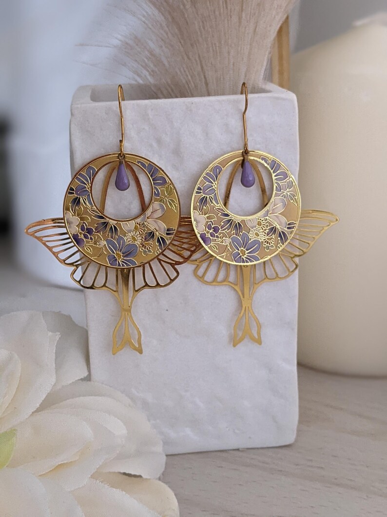 May include: Gold tone earrings with a floral design and a purple teardrop accent. The earrings are shaped like butterflies with delicate wings.