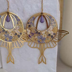 May include: A pair of gold earrings with a delicate floral design and a purple enamel accent. The earrings are shaped like a crescent moon with a dangling tail.