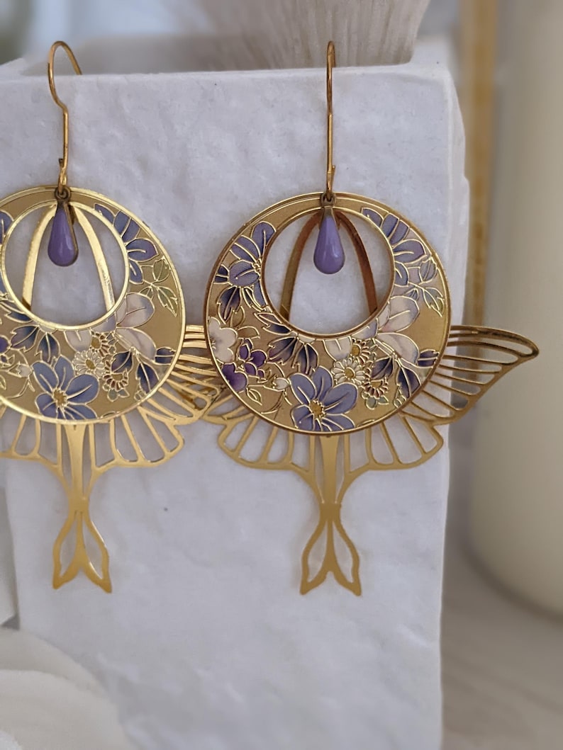 May include: A pair of gold earrings with a delicate floral design and a purple enamel accent. The earrings are shaped like a crescent moon with a dangling tail.