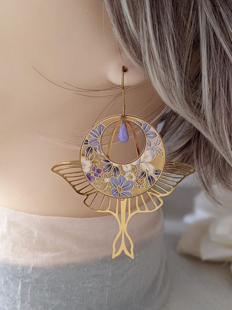 May include: Gold earrings with a floral design and a butterfly shape. The earrings are decorated with purple and white flowers on a gold background.