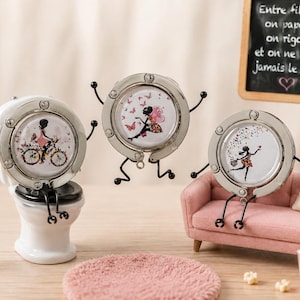 Peut inclure: Trois porte-sacs circulaires en métal argenté avec des inserts décoratifs illustrant des silhouettes. Un porte-sac est posé sur des toilettes miniatures blanches, un est suspendu en l'air et un est posé sur un canapé rose miniature.