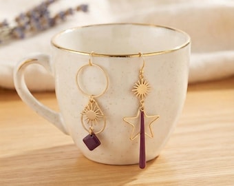 May include: A pair of mismatched gold-tone earrings hanging on a cream-colored ceramic mug. One earring features a circular hoop with a sunburst charm and a small purple square pendant. The other earring displays a sunburst charm above a star outline.