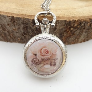 Peut inclure: Pendentif montre de poche argenté avec une chaîne. Le cadran de la montre présente une rose rose tendre et un motif de clés antiques. La montre ronde a une lunette décorative et une tige de remontoir. Fond en bois.