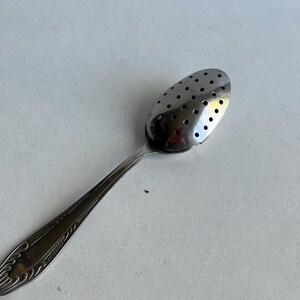 Può includere: Un cucchiaino infusore per tè argentato con una ciotola ovale perforata e un lungo manico decorato. Il cucchiaino è realizzato in metallo lucido ed è progettato per l'infusione di tè sfuso. Il manico presenta un design ornato e inciso.