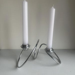 May include: A modern, silver metal candle holder with two white taper candles. The holder has a unique, looping design. The candles are approximately 25 cm tall.