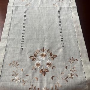 以下が含まれることがあります： 両端に茶色と白の複雑な花柄の刺繍が施された長方形のベージュのテーブルランナー。中央には縦に並んだ透かし彫りのヘムステッチのディテールがあり、縁には装飾的なボーダーがあしらわれています。