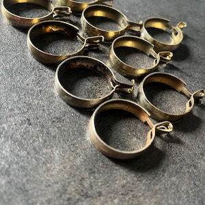 Puede incluir: Un conjunto de ocho anillas circulares para cortinas hechas de latón, cada una con un pequeño ojal integrado en el lateral. Los anillos de metal presentan un acabado dorado desgastado y deslustrado con signos visibles de edad y oxidación.