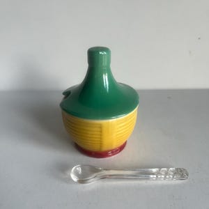 May include: A ceramic condiment jar with a green lid and yellow body, resting on a red base. A small, clear spoon is next to the jar. The jar has a small opening for the spoon. The jar is likely for salt or sugar.