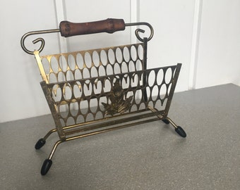May include: A gold metal magazine rack with a wooden handle and black feet. The rack has a decorative leaf design.