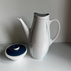Könnte beinhalten: Weiße Keramik-Kaffeekanne mit marineblauem Deckel. Die Kanne hat einen gebogenen Griff und einen Ausguss. Sie ist mit dünnen blauen und schwarzen Linien verziert. Der Deckel ist rund mit einem marineblauen Oberteil und einem weißen Rand.