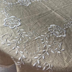 Puede incluir: Un mantel cuadrado de color beige con motivos florales bordados en blanco a lo largo del borde. El diseño incluye flores estilizadas, enredaderas sinuosas y pequeños detalles circulares de ojal cosidos en la tela, creando un marco.
