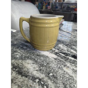 May include: A small, yellow ceramic pitcher with a handle and a spout. The pitcher has a barrel-like design with horizontal ridges. The interior is white. The pitcher is sitting on a speckled gray and white surface.