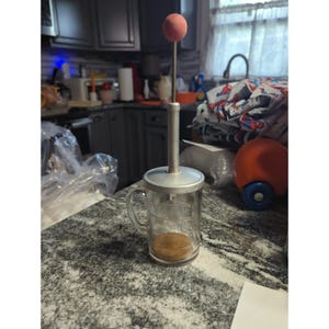 May include: A clear glass measuring cup with a silver-toned metal lid and a vertical plunger mechanism topped with a small red sphere. The glass features embossed measurement markings on the side and a handle, with a brown substance at the base.