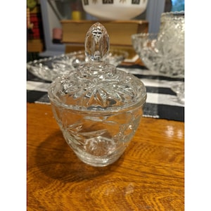 Può includere: Una bomboniera in vetro trasparente e vintage con coperchio. Il piatto ha una base rotonda e un coperchio con manico decorativo. Il vetro è inciso con un motivo floreale. Il piatto è su una superficie di legno.