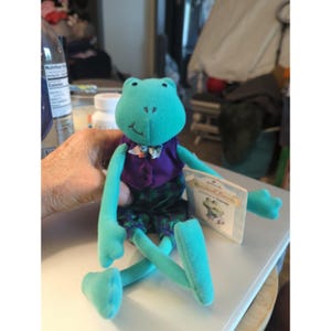 May include: A plush frog toy with bright teal skin, wearing a purple vest over a floral bowtie and dark green plaid pants. The frog has a stitched mouth and eyes, and it is attached to a rectangular Hallmark tag that reads Hallmark Keepsake Friends.