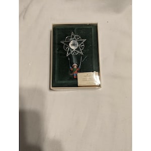 Pode incluir: Um enfeite de Natal Hallmark de 1981, apresentado na caixa original. O enfeite apresenta um design em forma de estrela com uma pequena figura em um balanço. A figura usa roupas vermelhas, azuis e verdes, em um fundo verde.