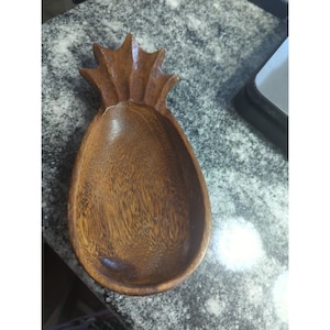 Op de afbeelding: Een houten serveerschaal in de vorm van een ananas. De schaal is bruin met een natuurlijk houtnerfpatroon. De bovenkant van de schaal is gevormd als een ananaskroon en de kom is ovaal van vorm.