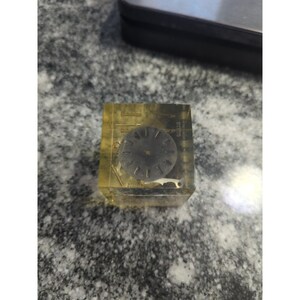 May include: A clear, yellow resin cube encasing watch parts, including a dark grey watch face with hour markers. The cube contains small metal components and is set against a grey speckled surface. The watch face is approximately 2.5 cm in diameter.