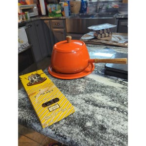 May include: A bright orange fondue pot with a matching lid and a long wooden handle, resting on a circular orange base. Beside it lies a rectangular yellow box with diagonal stripes, labeled Fondue Fork Set, featuring an illustration of forks.