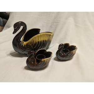 Puede incluir: Un conjunto de tres figuras de cisnes de cerámica en tonos marrón y amarillo. El cisne más grande es una maceta, y los más pequeños son candelabros. Los cisnes tienen un acabado brillante y están sobre una superficie blanca.