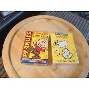 May include: Two decks of Peanuts-themed playing cards. One deck features Charlie Brown on a red background, the other features Snoopy on a yellow background. Both decks are rectangular and have the words "Playing Cards" on them.