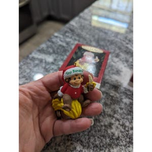 May include: A Christmas ornament featuring a monkey wearing a Santa hat with the text "Top Banana." The monkey is holding a banana and sitting on a yellow banana peel. The ornament is red, brown, and yellow.