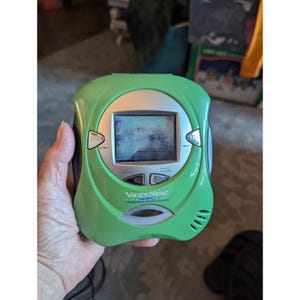May include: A lime green VideoNow Color personal video player featuring a small square screen, silver control buttons labeled Prev, Next, Stop, and Play/Pause, and a speaker grille. The device has the text VideoNow Color Personal Video Player printed.