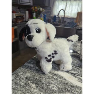 Può includere: Un peluche a forma di cane dalmata, principalmente bianco con macchie e dettagli neri. Il giocattolo ha un orecchio, un naso e dettagli degli occhi neri. Il cane è su una superficie maculata.