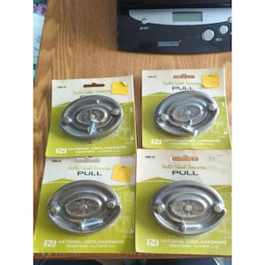 May include: Four vintage cabinet pulls on their original packaging. The pulls are oval-shaped with a decorative center and a brass finish. The packaging reads "Quality Cabinet Accessories" and "PULL."