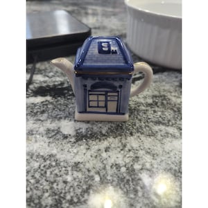 Può includere: Una piccola teiera in ceramica a forma di casa blu, con porta e finestre bianche. Il tetto è blu con una piccola finestra quadrata. Il beccuccio e il manico sono bianchi con finiture dorate. Su una superficie grigia macchiettata.