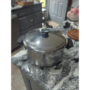 Puede incluir: Una olla de acero inoxidable con tapa a juego y dos asas laterales negras. La olla presenta un acabado metálico pulido y reflectante, con un pomo negro circular centrado en la tapa. El diseño incluye un cuerpo cilíndrico y una tapa plana.