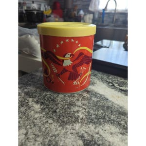 Puede incluir: Un recipiente cilíndrico rojo con tapa amarilla. El recipiente presenta un diseño de águila con las alas extendidas, sosteniendo flechas y una pancarta. El diseño es en rojo, amarillo y blanco. Estrellas están encima del águila.