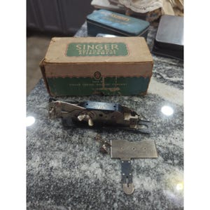May include: Vintage Singer buttonhole attachment kit in its original box. The box is tan with green and gold lettering. The attachment is silver and black, with a small metal plate. The box reads "Singer Buttonhole Attachment".