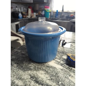 Może przedstawiać: Niebieski ceramiczny wolnowar z przezroczystą pokrywką. Wolnowar ma pionowe żebrowane detale i przezroczysty uchwyt na pokrywce. Stoi na nakrapianym blacie.