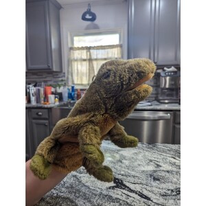 May include: A plush, brown and green hand puppet shaped like a lizard or dinosaur. The puppet has a textured, furry appearance with a lighter-colored mouth and darker green feet. The puppet is being held up by a person's hand.