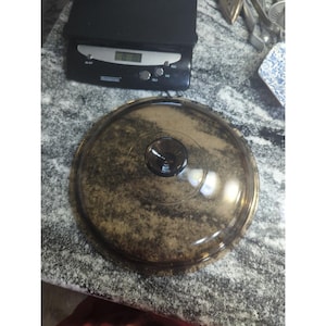 Puede incluir: Una tapa redonda transparente, moteada en marrón y beige, con un pomo marrón oscuro en el centro. La tapa está hecha de un material transparente. Utensilios de cocina.