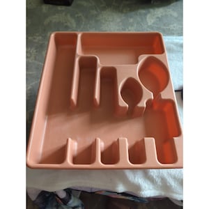 May include: A peach-colored plastic cutlery tray featuring a rectangular frame with multiple molded compartments of varying shapes and sizes, including a long side slot, a horizontal top section, and several smaller vertical slots for utensils.