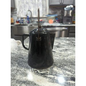 May include: Black enamelware coffee pot with white speckles. The pot has a spout, a handle, and a lid with a handle. The handle is made of metal. The pot is sitting on a granite countertop. Kitchenware.
