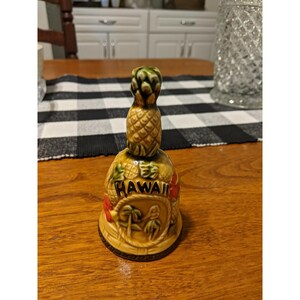 Puede incluir: Una campana de cerámica con forma de piña, con la parte superior verde y el cuerpo marrón. La palabra "Hawaii" está escrita en la campana, junto con imágenes de palmeras y flores. La campana está sobre una mesa de madera.