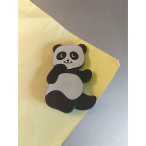 May include: A panda-shaped pin featuring a black and white color scheme. The design depicts a seated panda with black ears, black eye patches, black limbs, and a white face and belly. The panda has a simple facial expression with small eyes and a mouth.