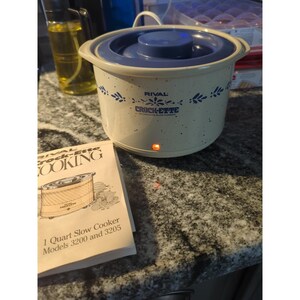 May include: A Rival Crock-ette 1-quart slow cooker with a blue lid and floral design. The slow cooker is cream-colored with blue speckles and a red power indicator light. A manual is next to the slow cooker.