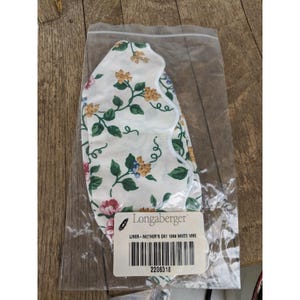 Può includere: Un rivestimento in tessuto bianco con un motivo floreale di foglie verdi e fiori gialli, rosa e blu, confezionato in un sacchetto di plastica trasparente. Il rivestimento è etichettato "Longaberger" e "Liner - Mother's Day 1999 White Vine."