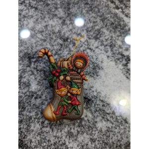 May include: A ceramic Christmas stocking ornament featuring elves, a child in a hood, and a candy cane. The stocking is tan, brown, and green, with red and green accents. A gold string is attached for hanging.