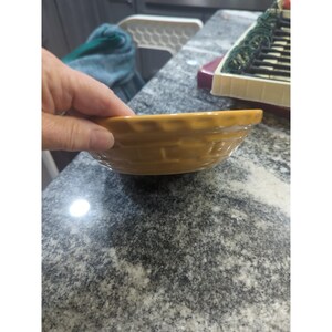 May include: A small, round, yellow-brown ceramic bowl with a basket weave pattern around the sides. The bowl is held by a hand, and the background is a speckled countertop.