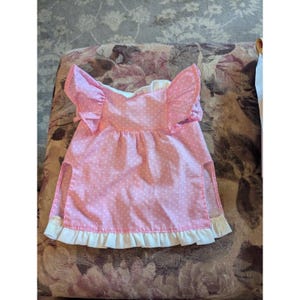 May include: A pink doll dress with white polka dots and ruffled sleeves. The dress has a white ruffled hem and a split on each side. The dress is made of a lightweight fabric.