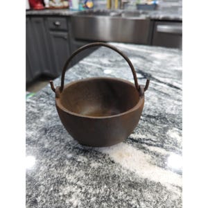 May include: A small, round, dark brown cast iron cauldron with a textured surface and a thin, arched metal handle attached to two side lugs. The bowl-shaped vessel features a deep interior and a sturdy, heavy-duty construction throughout.